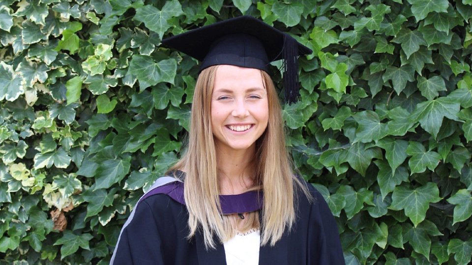 megan in graduation cap and gown posed in front of greenery on campus
